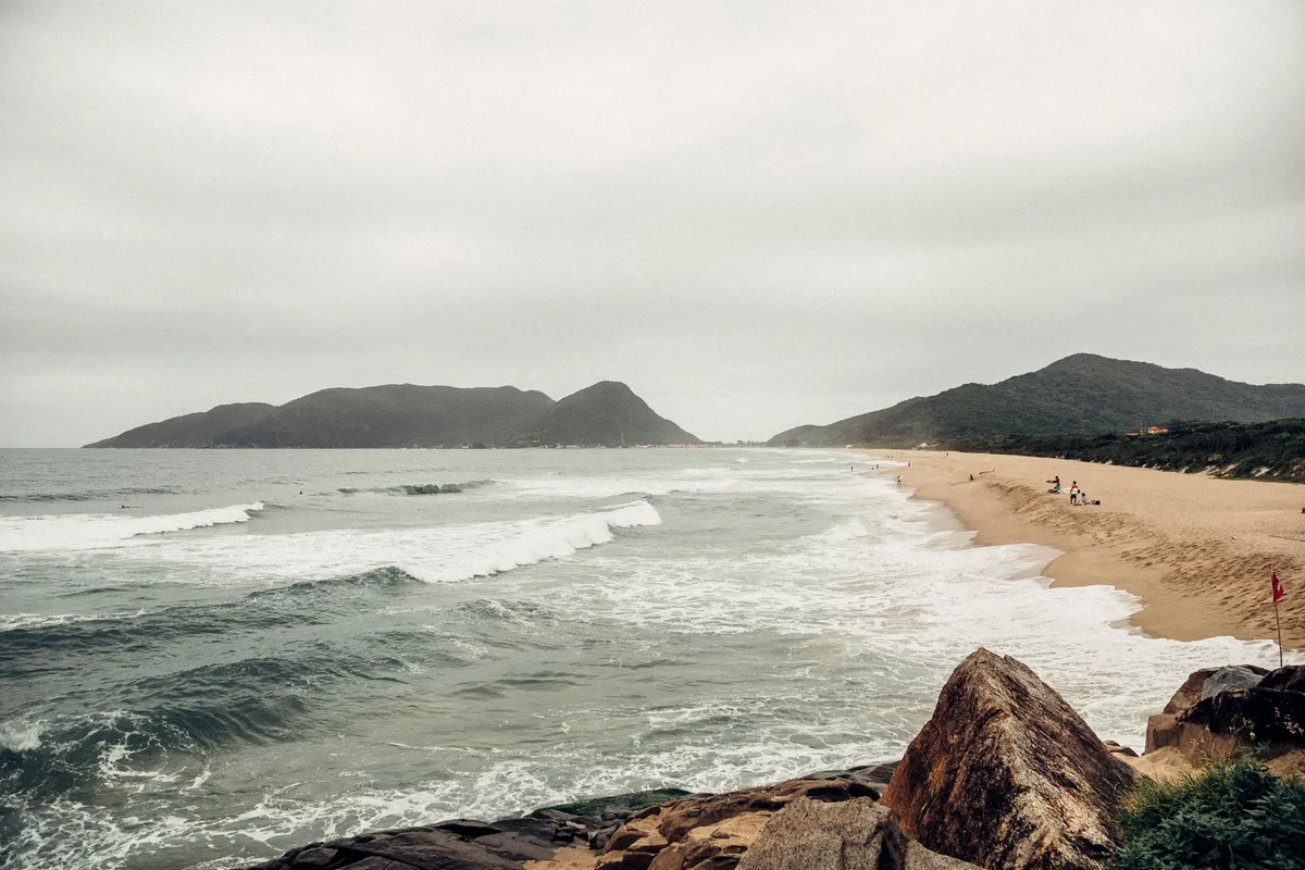 Praia urbana de Florianópolis com mar calmo e embarcações ao fundo