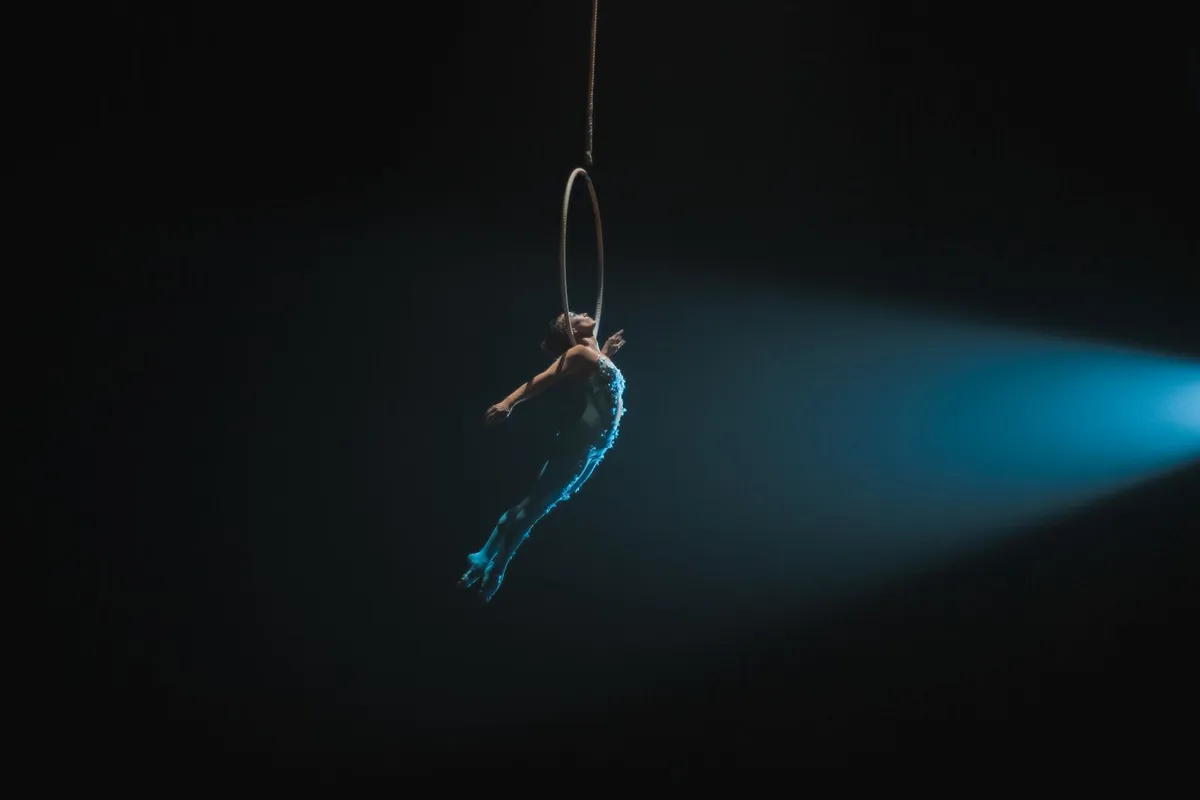 Artista de circo em acrobacia aérea no aro, iluminada por um feixe de luz azul