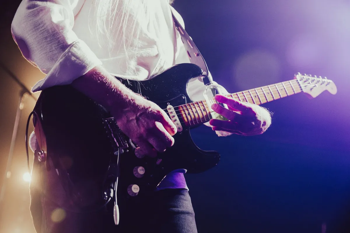 Close de guitarrista tocando guitarra elétrica preta sob luz roxa em show