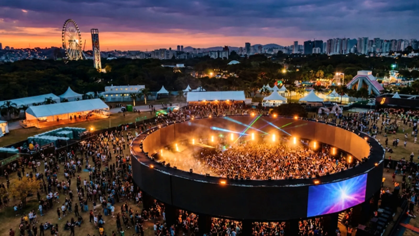 Vista aérea de festival de música eletrônica ao pôr do sol, com roda-gigante, tendas e palco circular lotado
